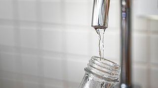 Ein Wasserhahn, aus dem klares Wasser in eine Glasflasche fließt. Der Wasserhahn ist aus glänzendem Metall.