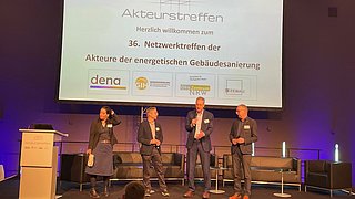 Vier Personen stehen auf einer Bühne vor einer Leinwand mit der Überschrift 'Akteurstreffen' und mehreren Logos darunter