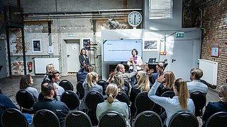 Zwei Personen halten einen Vortrag vor einem Publikum. Menschen im Publikum melden sich.
