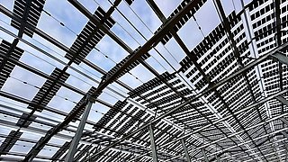 Blick nach oben auf ein Dach aus vielen rechteckigen Solarpaneelen, die von Metallstreben getragen werden.