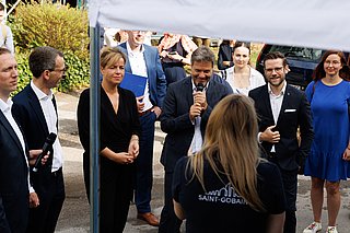 Eine Gruppe von Personen steht im Freien, einige halten Mikrofone. Ein Robert Habeck spricht in ein Mikrofon, während andere zuhören. Im Hintergrund sind Fahrzeuge und eine Menschenmenge sichtbar.