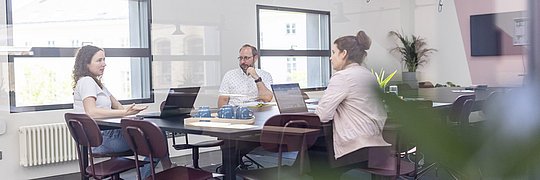 Drei Personen in einem modernen Büro, die an einem Tisch mit Laptops diskutieren.