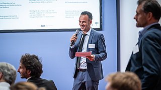 Mann im Anzug hält Mikrofon und rotes Blatt, spricht vor Publikum mit Präsentation im Hintergrund.