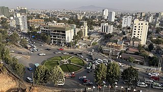 Luftaufnahme eines verkehrsreichen Kreisverkehrs mit Grünfläche und umliegenden mehrstöckigen Gebäuden in einer Stadt