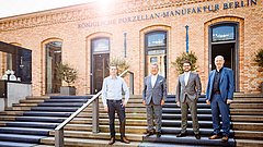 Vier Männer stehen auf einer Treppe vor der Königlichen Porzellan-Manufaktur Berlin. Die Männer tragen formelle Kleidung und stehen in einer Reihe. Im Hintergrund sind große Fenster und Pflanzen sichtbar.