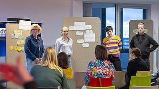 Vier Personen stehen vor Pinnwänden mit Zetteln, während mehrere Personen auf bunten Stühlen ihnen zuhören.