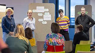 Vier Personen stehen vor Pinnwänden mit Zetteln, während mehrere Personen auf bunten Stühlen ihnen zuhören.