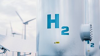 Großer Wasserstofftank mit der Aufschrift „H₂“, im Hintergrund Windräder und Solarpanel.