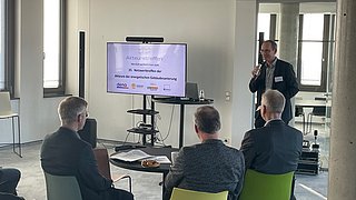 Vier Personen sitzen in einem Raum, eine steht mit Mikrofon vor einem Bildschirm mit Text zur 35. Netzwerkwerkstatt der Akteure der energetischen Gebäudesanierung.