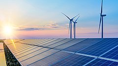 Solaranlage auf einem Dach mit mehreren Solarpanelen, im Hintergrund Windkraftanlagen und ein Sonnenuntergang.