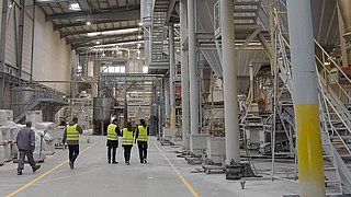  Vier Personen in Sicherheitskleidung gehen durch eine Werkhalle, in der eine Trockenanlage steht.