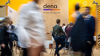 Messestand der dena mit Holzrückwand und gelben Seitenwänden, mehrere Personen im Gespräch, Schriftzug 'dena, Wir machen Energiewende'