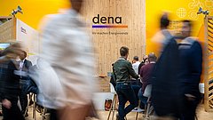Messestand der dena mit Holzrückwand und gelben Seitenwänden, mehrere Personen im Gespräch, Schriftzug 'dena, Wir machen Energiewende'