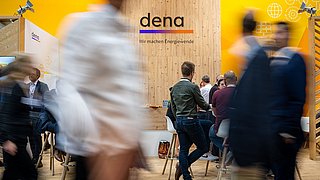 Messestand der dena mit Holzrückwand und gelben Seitenwänden, mehrere Personen im Gespräch, Schriftzug 'dena, Wir machen Energiewende'