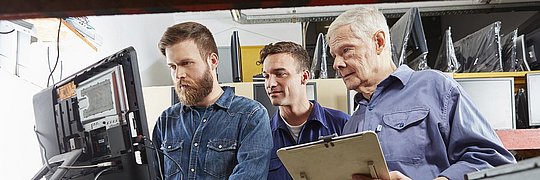 Drei Techniker stehen an einem Computerterminal in eienr Werkstatt.