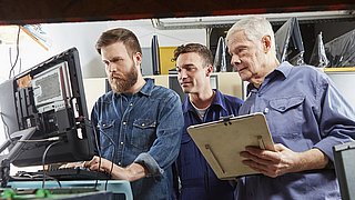 Drei Techniker stehen an einem Computerterminal in eienr Werkstatt.
