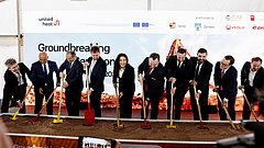 Gruppe von neun Personen in Anzügen bei einer symbolischen Spatenstich-Zeremonie unter einem Zelt vor einem Banner mit Logos und dem Schriftzug 'Groundbreaking Ceremony'