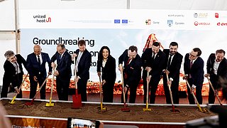 A group of nine people in suits at a symbolic ground-breaking ceremony under a marquee in front of a banner featuring logos and the words 'Groundbreaking Ceremony'
