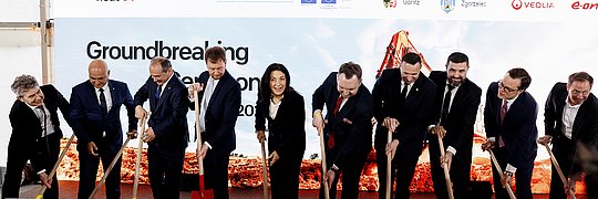 Gruppe von neun Personen in Anzügen bei einer symbolischen Spatenstich-Zeremonie unter einem Zelt vor einem Banner mit Logos und dem Schriftzug 'Groundbreaking Ceremony'