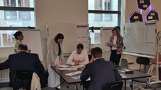 Mehrere Personen sitzen und stehen um einen Tisch in einem Büro mit Whiteboards und großen Fenstern