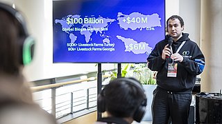 Person mit Mikrofon und schwarzem Hoodie hält Präsentation vor Bildschirm mit Weltkarte und Text zu globalem Biogasmarkt, Publikum mit Kopfhörern im Vordergrund