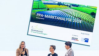 Drei Personen stehen an einem Tisch, im Hintergrund eine Präsentation mit dem Titel 'PPA-MARKTANALYSE 2024'. Darunter steht 'Marktorientierte Erneuerbare Energien' und das Datum 'August 2025'.
