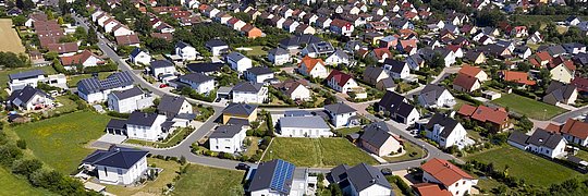 Luftaufnahme eines Wohngebiets mit vielen Einfamilienhäusern, grünen Gärten und einer Straße im Vordergrund.