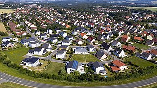 Luftaufnahme eines Wohngebiets mit vielen Einfamilienhäusern, grünen Gärten und einer Straße im Vordergrund.