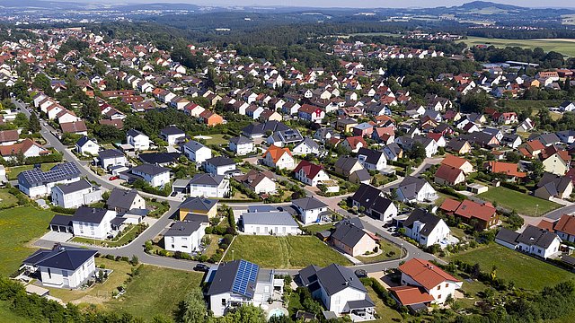 Luftaufnahme eines Wohngebiets mit vielen Einfamilienhäusern, grünen Gärten und einer Straße im Vordergrund.