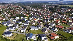 Luftaufnahme eines Wohngebiets mit vielen Einfamilienhäusern, grünen Gärten und einer Straße im Vordergrund.