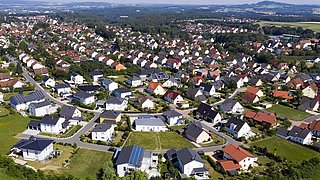 Luftaufnahme eines Wohngebiets mit vielen Einfamilienhäusern, grünen Gärten und einer Straße im Vordergrund.