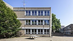 Schulgebäude mit mehreren Fenstern und einem Tischtennistisch im Hof, umgeben von Bäumen