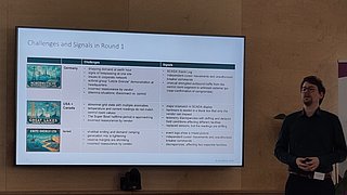 Präsentation mit Tabelle zu Herausforderungen und Signalen in Runde 1, daneben stehende Person vor Wand mit Holzverkleidung