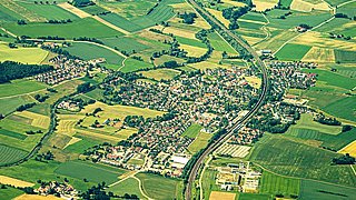 Luftaufnahme eines Dorfes mit umgebenden landwirtschaftlichen Feldern und einer Straße, die durch die Landschaft verläuft.