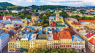 Eine bunte Stadtansicht mit historischen Gebäuden, vielen Bäumen und einer hügeligen, bewaldeten Landschaft im Hintergrund. Dominiert wird das Bild von zwei Kirchen mit markanten Türmen.