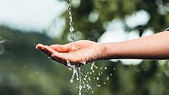 Eine Hand hält Wasser, das in einem dünnen Strahl herabfließt. Wassertropfen spritzen in alle Richtungen. Unscharfer Hintergrund mit grünen Bäumen.
