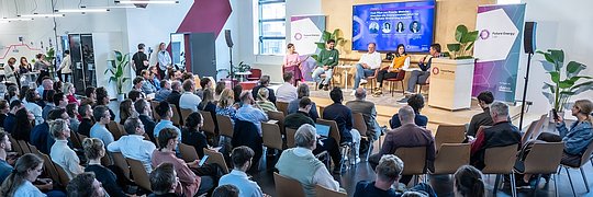 Großer Saal mit Publikum, das einer Podiumsdiskussion mit mehreren Personen auf der Bühne folgt.
