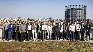 Gruppe von Menschen steht auf einem begrünten Dach mit Stadtpanorama und einem großen, runden Industriedenkmal im Hintergrund.