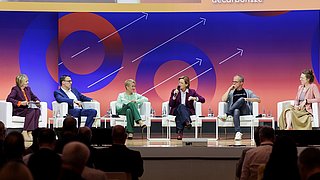 Sechs Personen sitzen auf weißen Sesseln auf einer Bühne vor einem orange-blauen Hintergrund mit Pfeil-Grafiken 