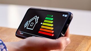 Person hält rosa Tasse und Smartphone mit Energieeffizienzdiagramm auf einem Holztisch.