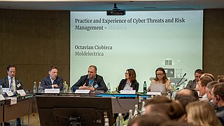 Mehrere Personen sitzen an einem langen Konferenztisch in einem Tagungsraum, eine Person spricht und gestikuliert, im Hintergrund eine Projektionswand mit dem Text 'Practice and Experience of Cyber Threats and Risk Management - Moldova' und 'Octavian Ciobirca Moldelectrica'