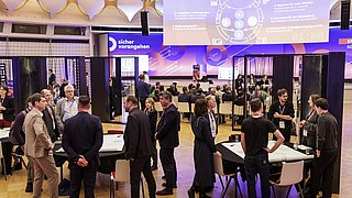 Mehrere Personen stehen und sitzen in einem großen, modernen Veranstaltungsraum mit Tischen und Stühlen, im Hintergrund eine große Projektionsfläche mit Text und Grafiken.