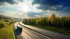 Green truck driving on the asphalt highway between deciduous forest in autumn colors under the radiant sun and dramatic clouds. View from above.