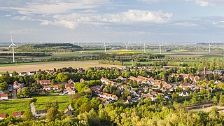 Luftaufnahme einer ländlichen Gegend mit Wohnhäusern, Feldern, Waldgebiet und Windkraftanlagen unter einem bewölkten Himmel.
