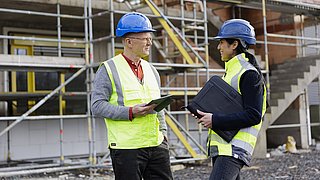 Zwei Personen in Sicherheitswesten und blauen Helmen stehen auf einer Baustelle. Eine Person hält ein Tablet, die andere eine schwarze Aktentasche. Im Hintergrund sind Gerüste und eine Außenwand sichtbar.
