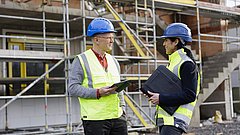 Ein Energieeffizienz-Experte und eine Energieeffizienz-Expertin stehen auf einer Baustelle vor einem Gerüst und unterhalten sich.  