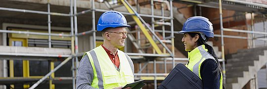 Ein Energieeffizienz-Experte und eine Energieeffizienz-Expertin stehen auf einer Baustelle vor einem Gerüst und unterhalten sich.