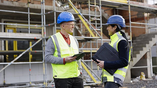 Zwei Personen in Sicherheitswesten und blauen Helmen stehen auf einer Baustelle. Eine Person hält ein Tablet, die andere eine schwarze Aktentasche. Im Hintergrund sind Gerüste und eine Außenwand sichtbar.