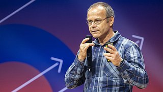 Mann in kariertem Hemd und beiger Hose hält Mikrofon und Präsentationsfernbedienung vor farbigem Hintergrund mit Pfeilgrafiken.