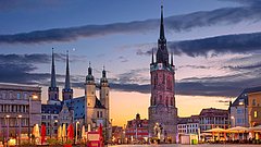 Stadtansicht von Halle mit mehreren Türmen, darunter ein hoher, spitz zulaufender Turm und drei weitere mit Zinnen. Abendhimmel mit Wolken und Mond.